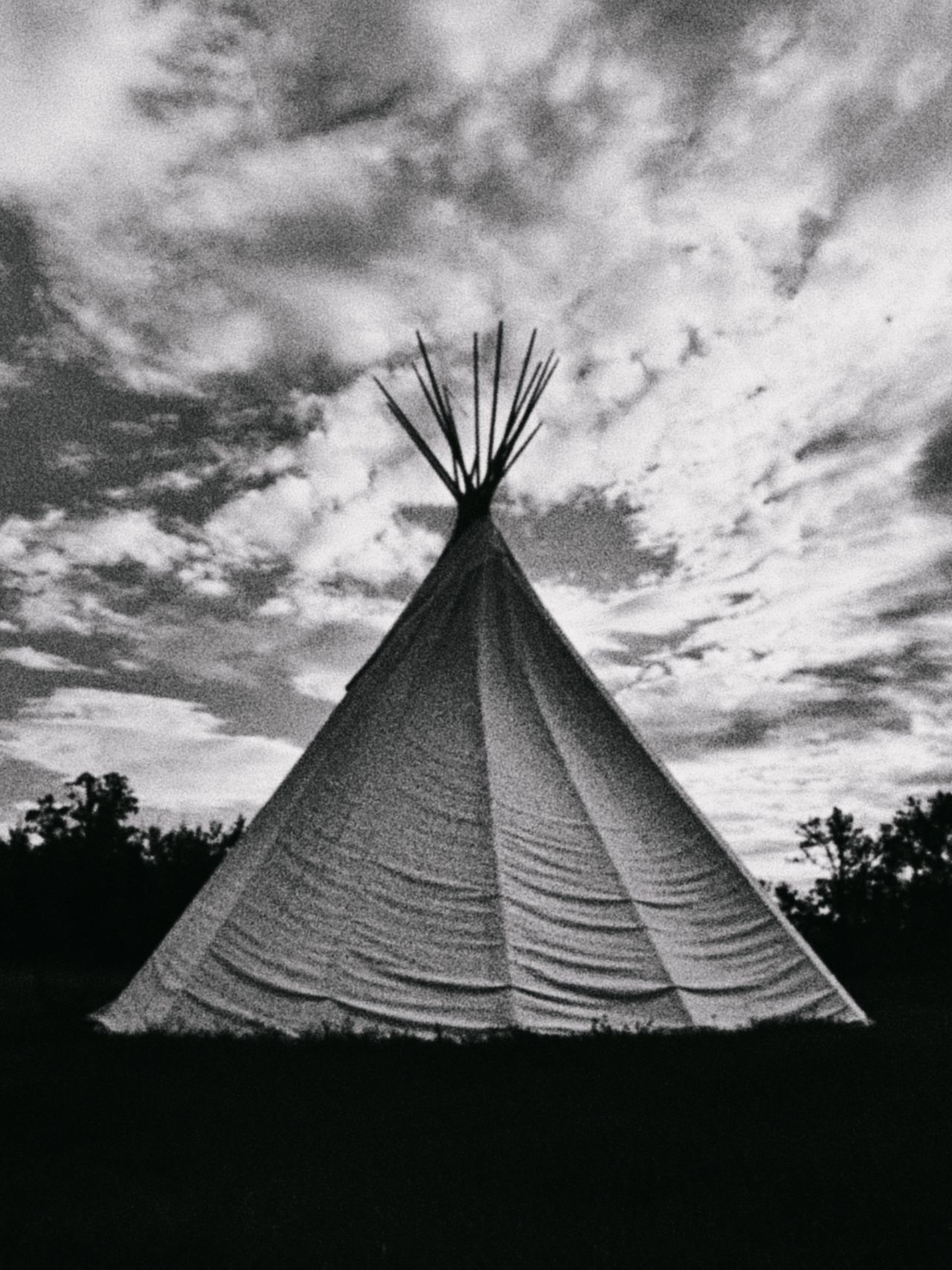 Black and white grainy high contrast photo of native american teepee in a dark field with cloudy sky