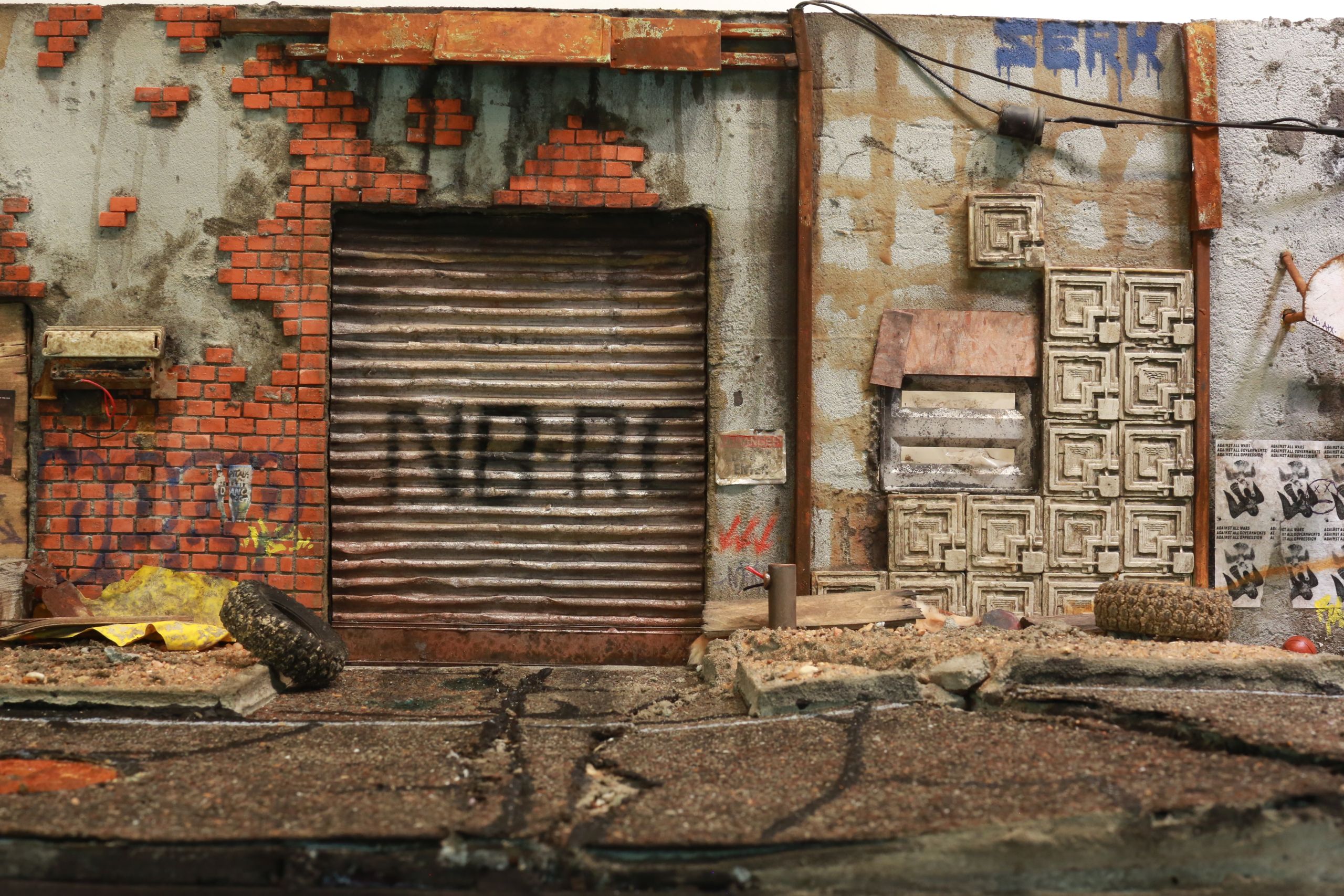 1/24-1/18 scale miniature diorama of a bombed out post apocalyptic inner city street alley way, with corrugated steel auto garage bay door, graffiti mural, and Frank Lloyd Wright Ennis House tiles