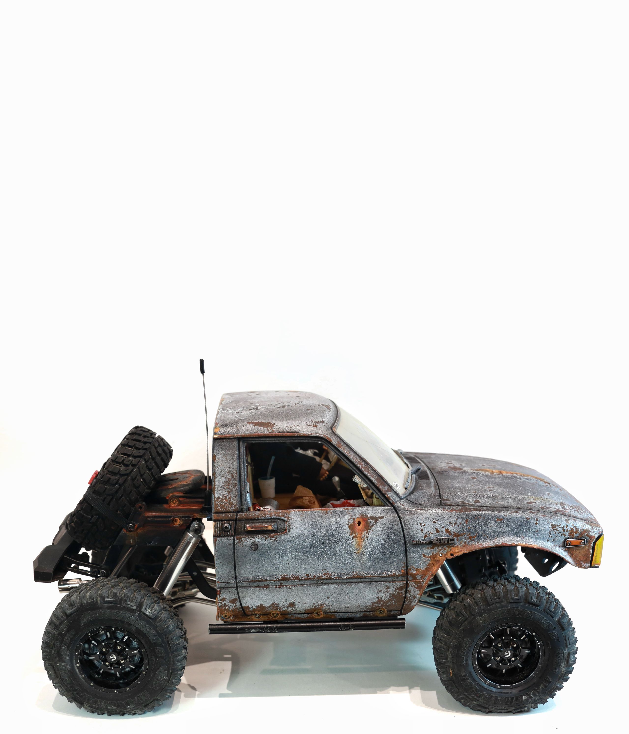 An old Toyota Hilux cab-only "beater" pickup truck working RC vehicle in 1/10 scale, with a gooseneck trailer hitch platform and carrying a spare wheel. Featuring a custom interior with action figure driver, handmade accessories, and faux leather bench seat.