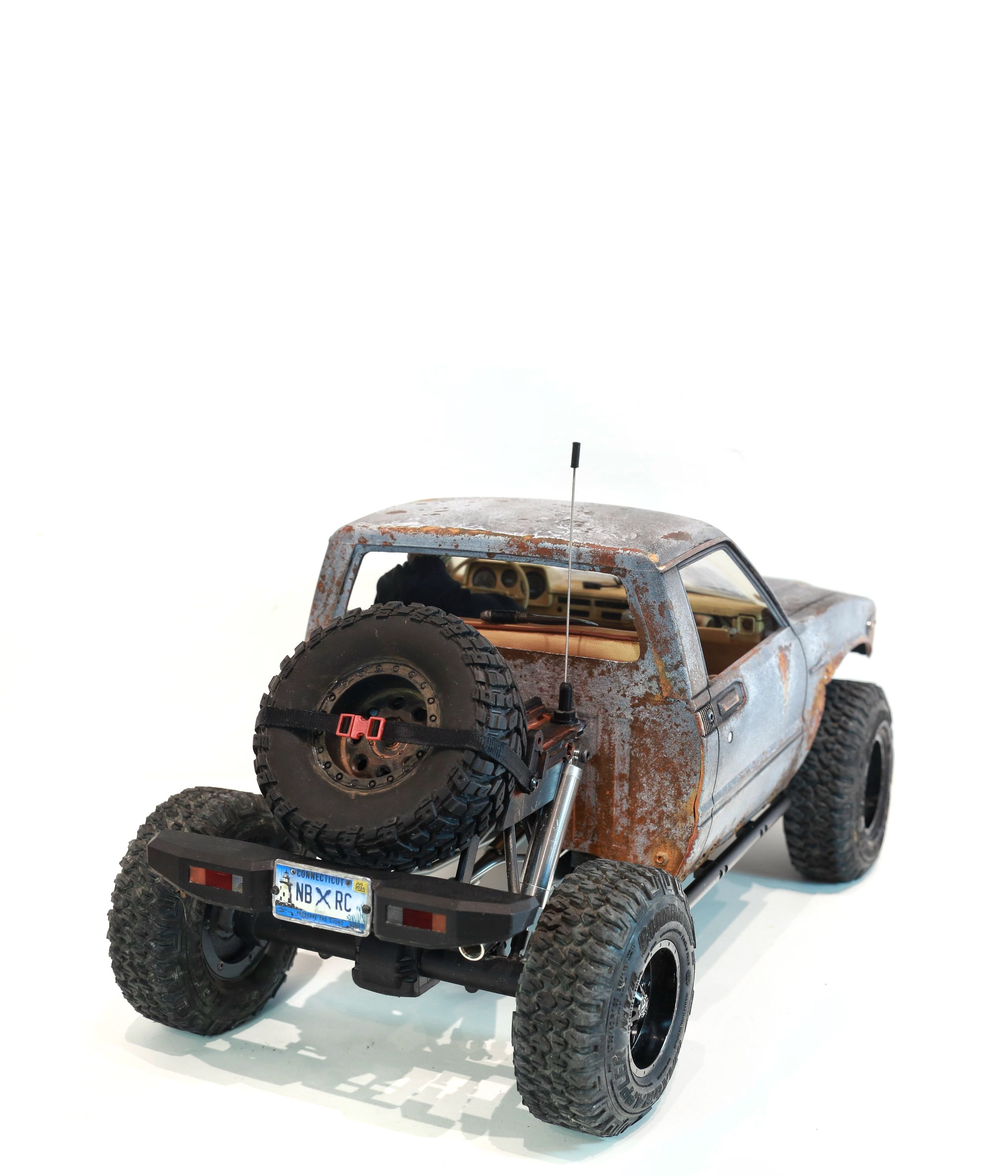 An old Toyota Hilux cab-only "beater" pickup truck working RC vehicle in 1/10 scale, with a gooseneck trailer hitch platform and carrying a spare wheel. Featuring a custom interior with action figure driver, handmade accessories, and faux leather bench seat.