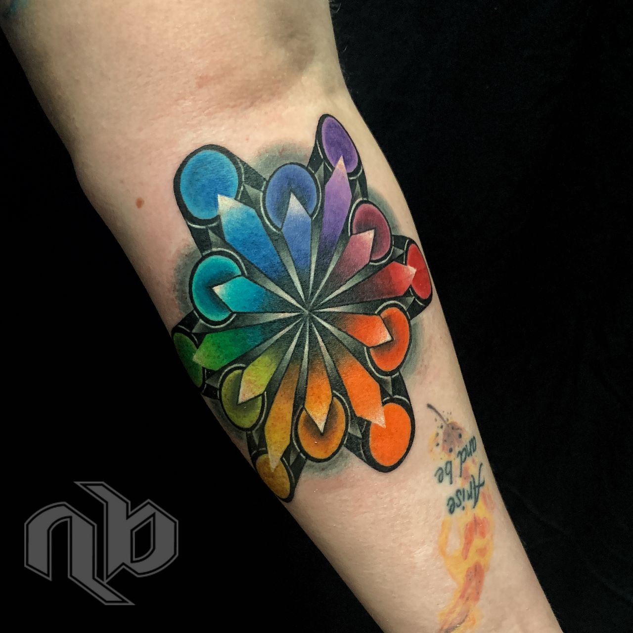Close view of a small arm tattoo with rainbow teardrops and circles radiating from a central point within bold black geometric outlines.