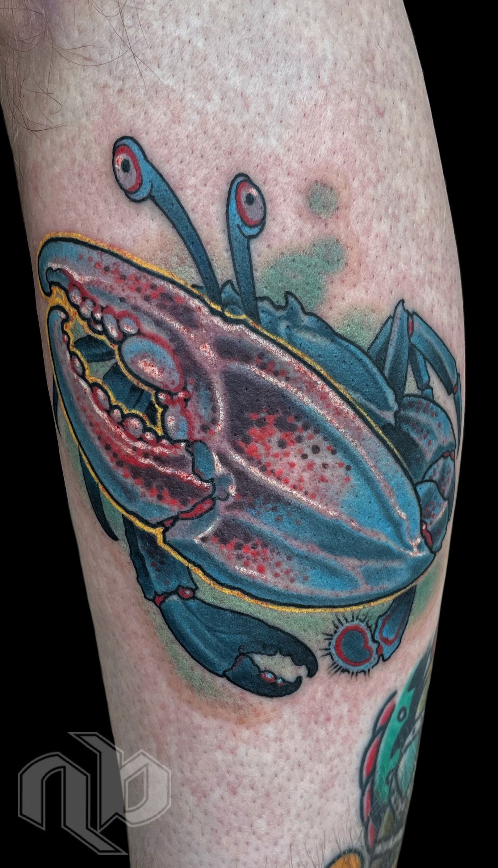 Close view of a blue crab tattoo on a leg, with red-speckled claws, raised eyestalks, and yellow outlines.