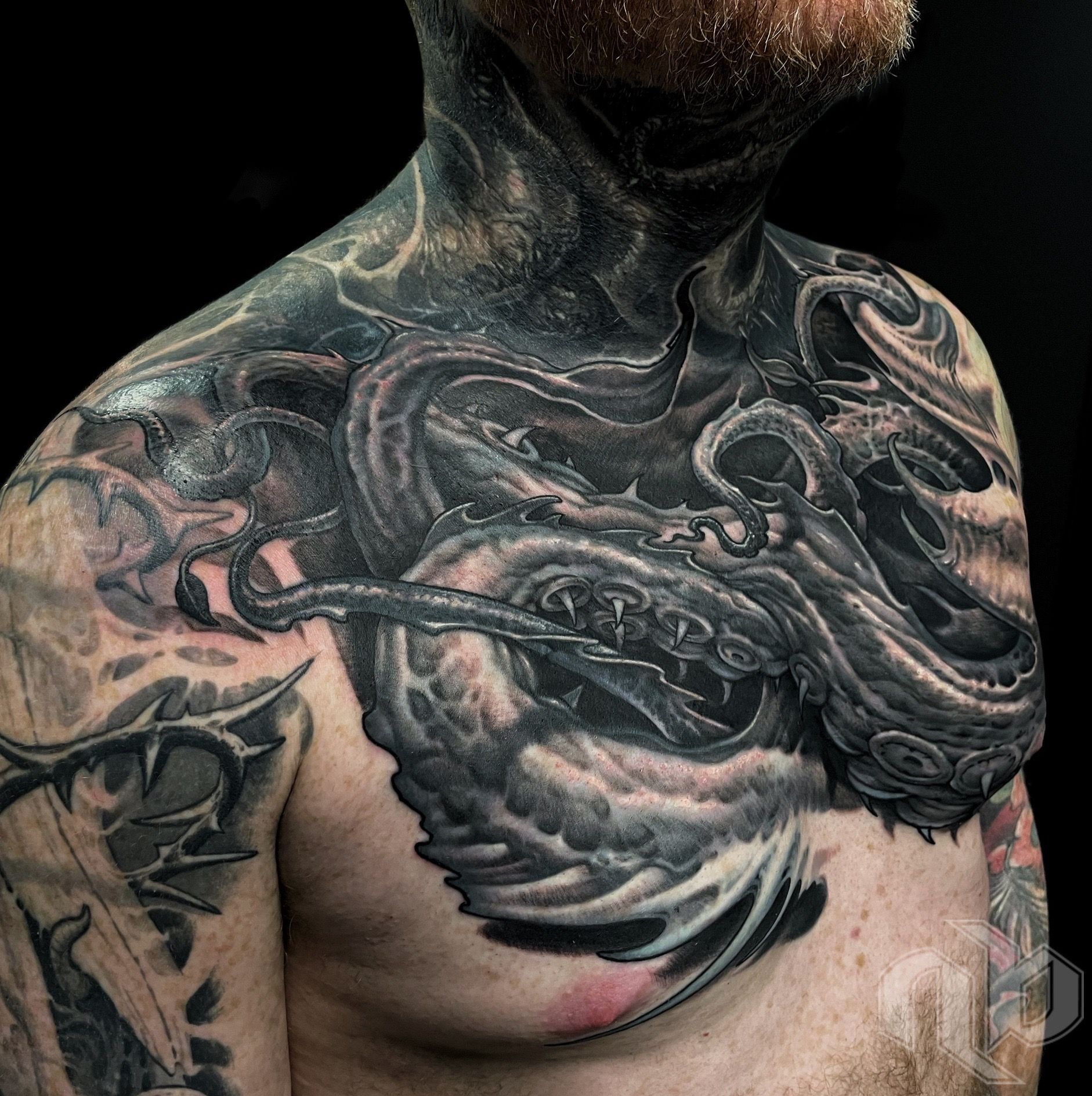 Close view of a black-and-grey chest tattoo featuring a fanged, tentacled creature extending across the shoulders and neck.