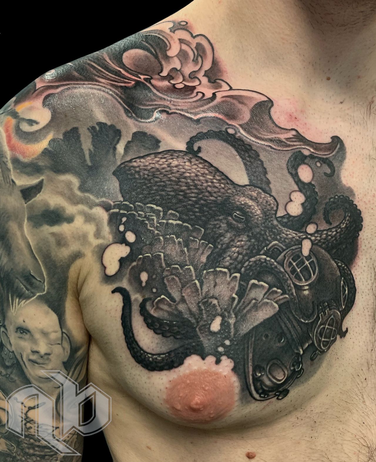 Close view of a black-and-grey chest tattoo depicting an octopus entwined around a deep-sea diver’s helmet amid stylized waves.