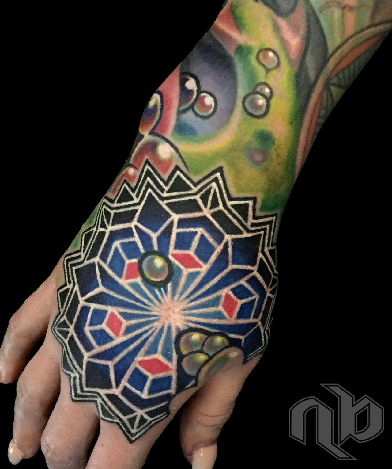 Close view of a hand bearing a blue geometric mandala tattoo with black edging, white outlines, red accents, and surrounding multicoloured designs.