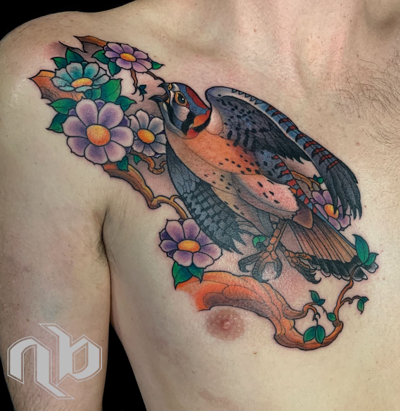 Close view of a colorful chest tattoo showing a kestrel bird perched among twisting branches, green leaves, and purple, blue, and orange flowers.