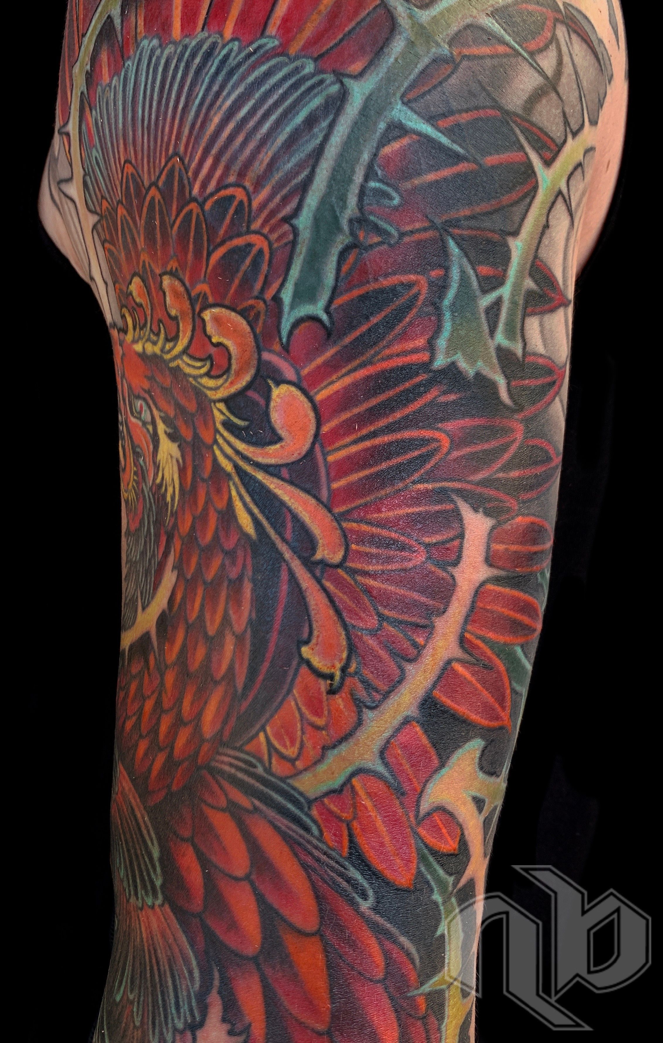 Close detail of a large arm tattoo showing a red-orange phoenix amid layered purple feathers and angular teal-and-yellow branch-like forms.