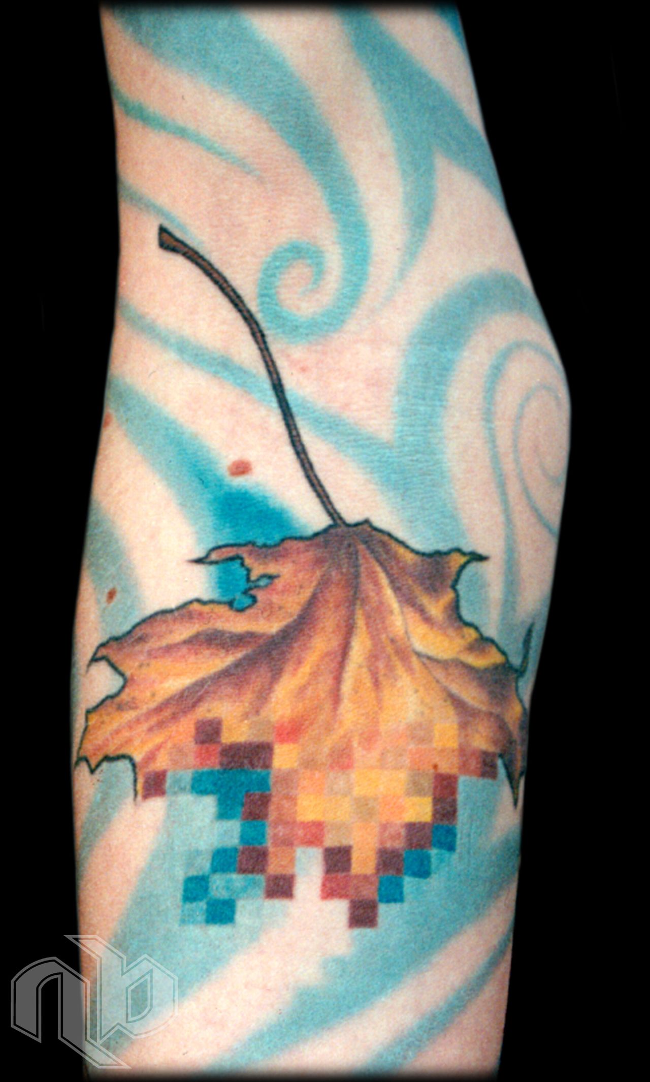 Close view of an arm tattoo showing an orange-brown leaf dissolving into colored pixels against swirling blue streaks.