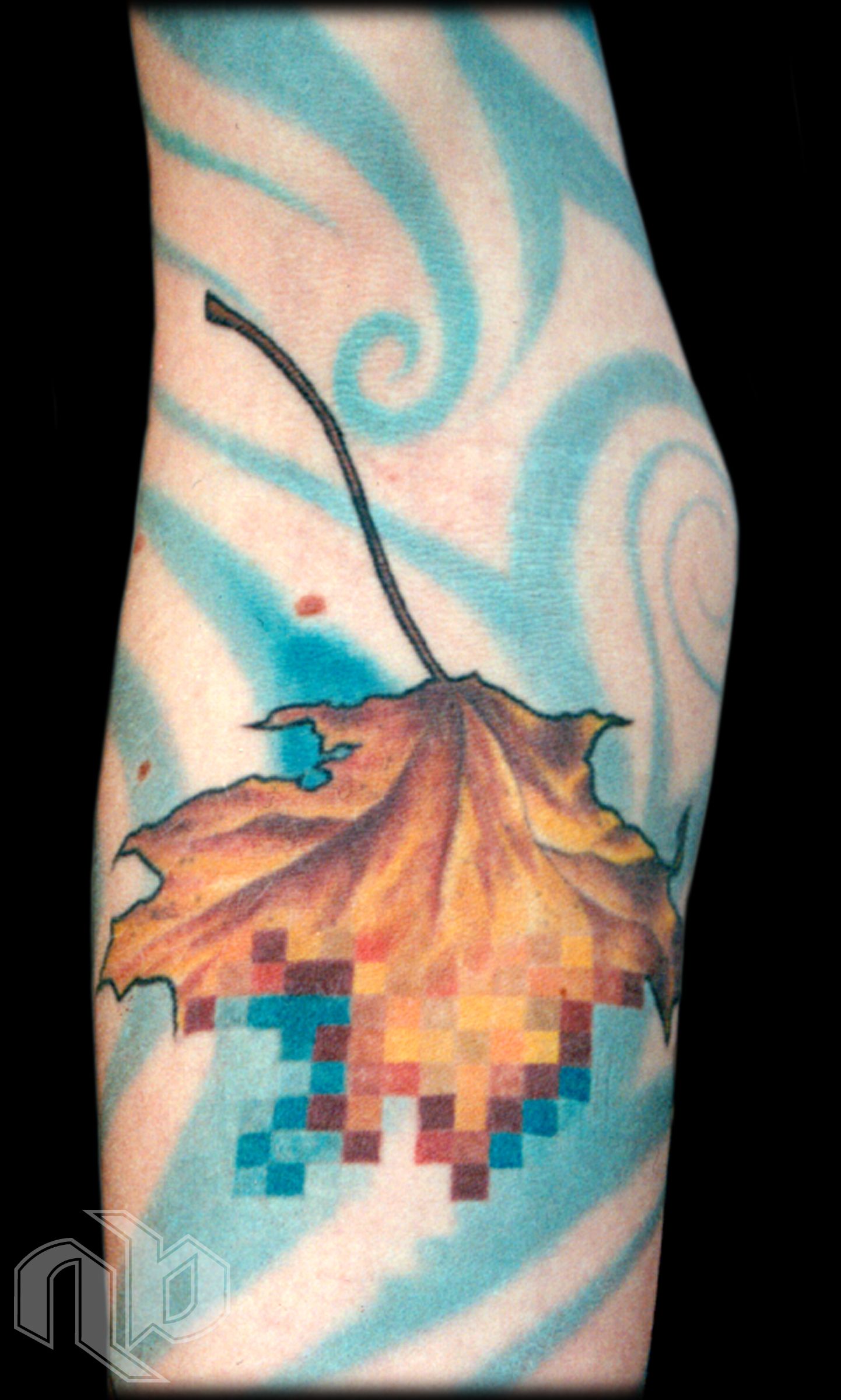 Close view of an arm tattoo showing an orange-brown leaf dissolving into colored pixels against swirling blue streaks.