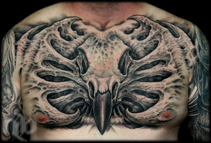 Black-and-grey chest tattoo featuring a central raven skull with a long beak, framed by symmetrical, perforated bone-like forms.