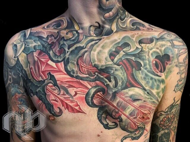 Close view of a torso with a large multicolored chest tattoo depicting a green bio-organic forms, red wings, clouds, and swirling forms.