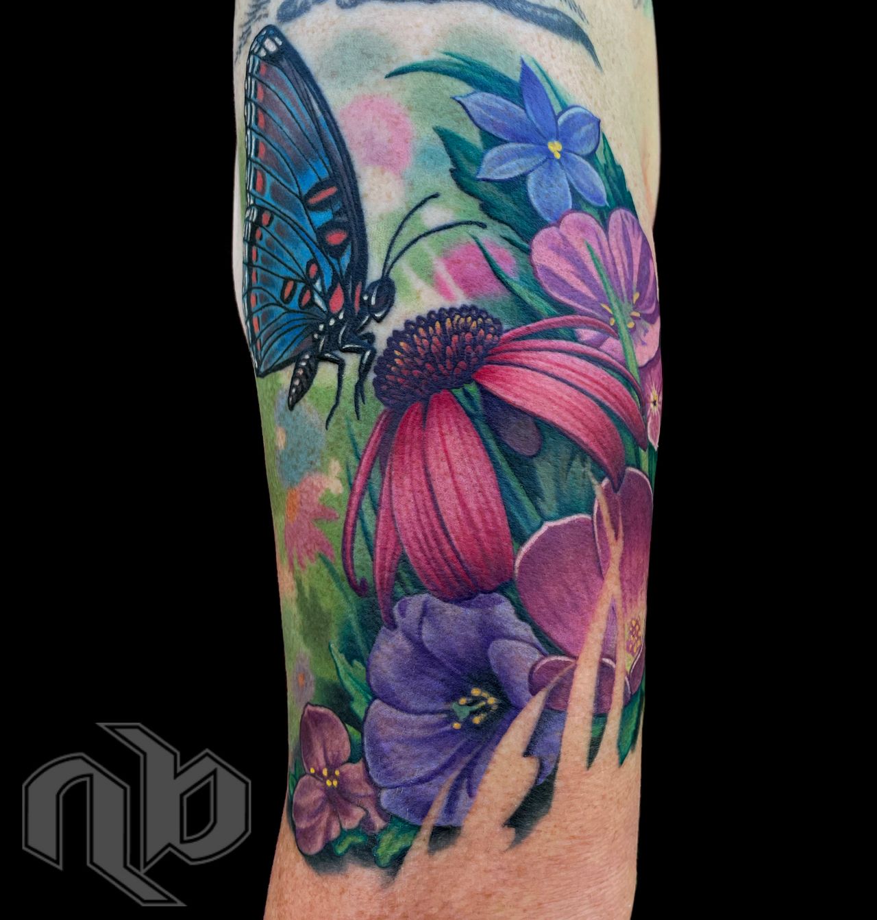 Close view of a colorful arm tattoo featuring a blue-and-black butterfly among pink, purple, and blue wildflowers and green leaves.